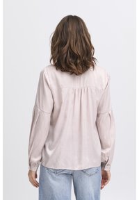 Long-sleeve light pink blouse with a slight sheen, pleated back detail, and buttoned cuffs, worn with high-waisted blue jeans.