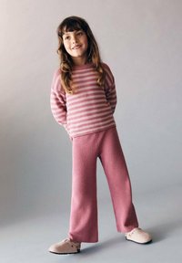 Pull en tricot rose à rayures avec des manches longues associé à un pantalon en tricot rose à jambe large. Pieds ornés de chaussures arrondies de couleur claire.