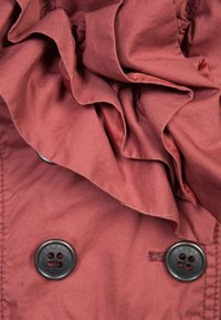 Rode stof met een gerimpelde kraag, voorzien van zwarte knopen en een gladde textuur. Het ontwerp bevat gelaagde stofdetails voor extra diepte.