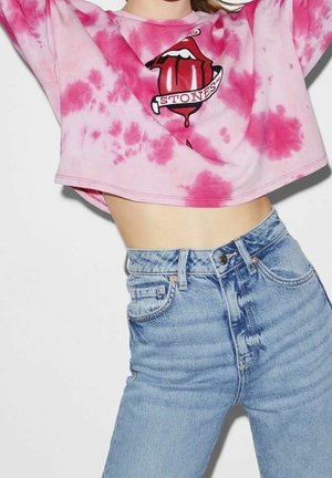Personne portant un sweat court tie-dye rose avec un motif graphique de langue et lèvres rouges et un jean en denim bleu clair taille haute.