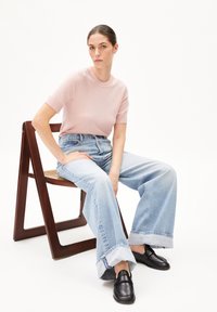 Light pink knitted short-sleeve top, high-waisted light blue denim jeans with wide legs, and black loafers, seated on a wooden chair.