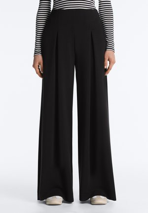Femme portant un pantalon noir taille haute à jambes larges, associé à des baskets blanches et un haut à manches longues rayé noir et blanc.