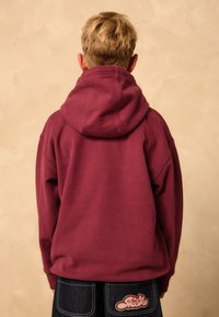 Felpa con cappuccio color bordeaux dal taglio oversize, caratterizzata da un cappuccio ampio e polsini a costine; abbinata a pantaloni in denim scuro con patch logo rosso.