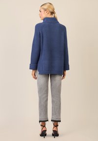 Blauer, gerippt gestrickter Pullover mit hohem Kragen und oversized Passform. Kombiniert mit hellgrauen, geraden Jeans mit fransigen Säumen.