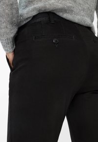 Pantaloni neri con una texture liscia, vestibilità su misura e una tasca posteriore con dettaglio a bottone. Indossati con un maglione grigio a pois.