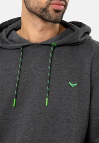 Homme barbu portant un sweat à capuche gris foncé avec des cordons de serrage vert fluo et un petit logo vert brodé sur la poitrine, tourné légèrement vers la droite.