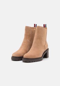 Botas de tobillo de ante marrón con un tacón grueso, paneles laterales elásticos y acentos de tab en contraste en rojo y azul en la parte trasera. Suela texturizada para mayor agarre.