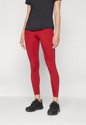 Personne portant des leggings rouges, des chaussures de sport noires et un t-shirt noir à manches courtes se tenant contre un fond gris clair uni.
