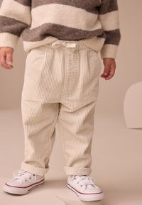 Pantalon en velours côtelé crème avec des rayures verticales texturées, doté d'une taille élastique et d'un nœud à l'avant, associé à des baskets blanches.