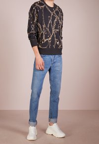 Felpa nera con motivo a catena dorata, scollo rotondo e polsini a costine, abbinata a jeans azzurri a culotte e sneaker bianche.