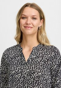 Schwarze Bluse mit weißem abstrakt Muster, V-Ausschnitt und knopflosem Verschluss. Hergestellt aus leichtem Stoff mit subtiler Textur.
