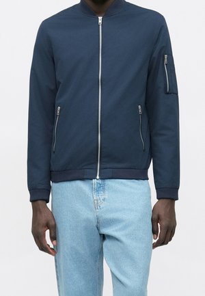 Mand iført navyblå bomberjakke med lynlåse og lyseblå jeans, stående mod hvid baggrund.