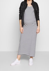 Schwangere Person, die eine schwarze Lederjacke, ein weiß-schwarz gestreiftes Kleid und weiße Sneakers trägt und vor einem schlichten Hintergrund steht.