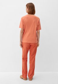 Kvinde med kort brunt hår står med ryggen til, iført en koralafsat kortærmet skjorte, matchende bukser og beige platformsko.