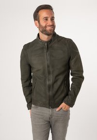 Grüne Lederjacke mit Stehkragen, Frontreißverschluss und zwei seitlichen Taschen. Strukturierte Oberfläche mit Nähdetails. Getragen über grauen Jeans.