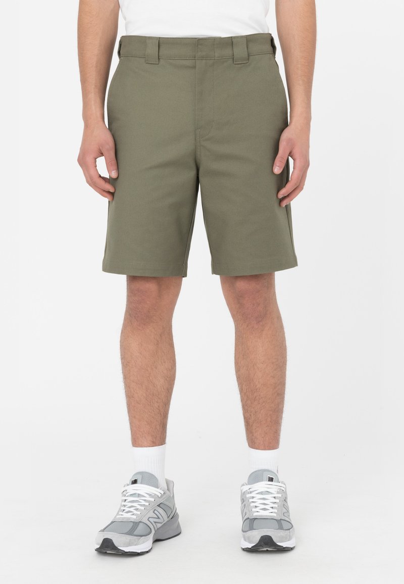 Pantaloni corti verde oliva con taglio dritto, realizzati in tessuto misto cotone, con passanti per cintura e tasche laterali. Abbinati a scarpe da ginnastica grigie.
