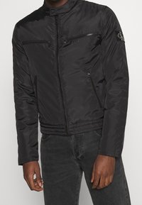 Giubbotto bomber nero realizzato in materiale liscio e leggero, dotato di due tasche sul petto con zip, polsini a costine e vita aderente.