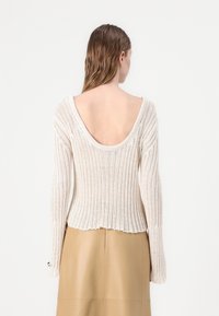 Maglione a maglia color crema con una texture a coste, scollatura ampia sul retro, maniche lunghe e lunghezza leggermente accorciata. Abbinato a una gonna color beige.