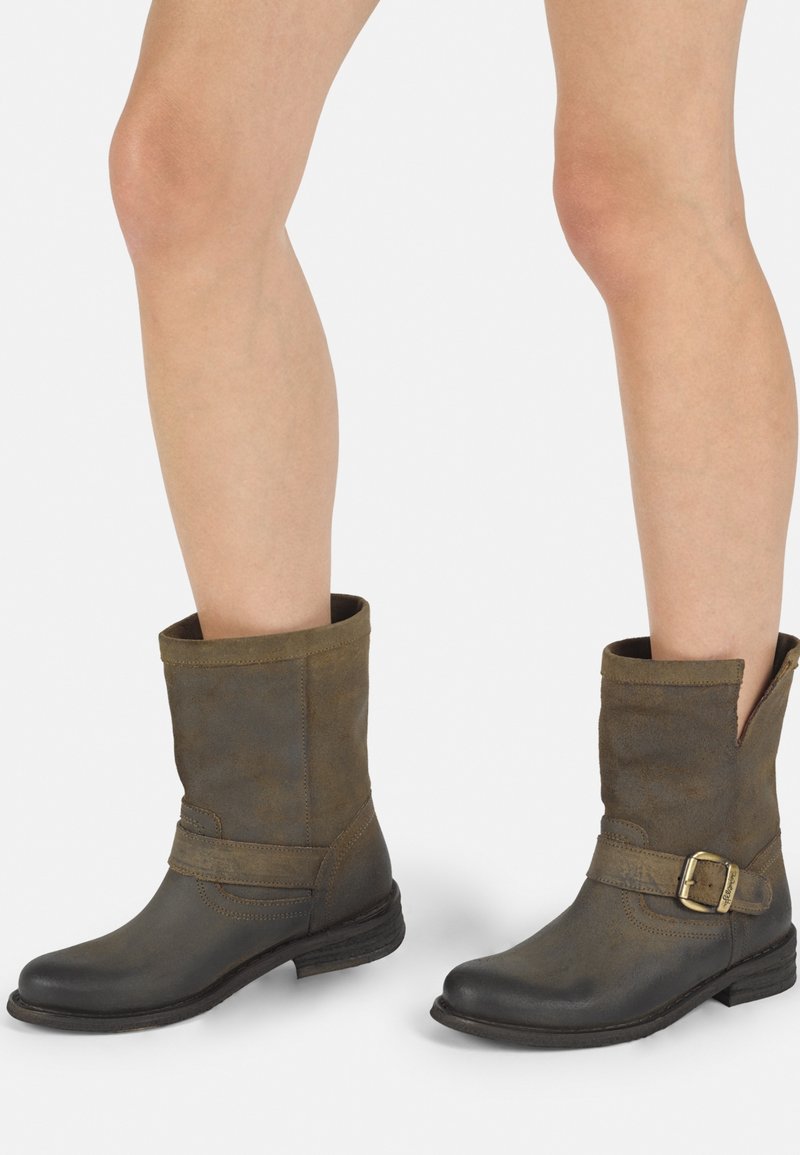 Botas de tobillo de cuero marrón con un acabado mate, que presentan una hebilla lateral, un eje ligeramente caído y un tacón bajo apilado.