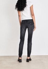 Jeans negros ajustados hechos de mezclilla, con cinco bolsillos y un lavado oscuro. La modelo lleva sandalias de tacón negras y una camisa blanca ajustada.