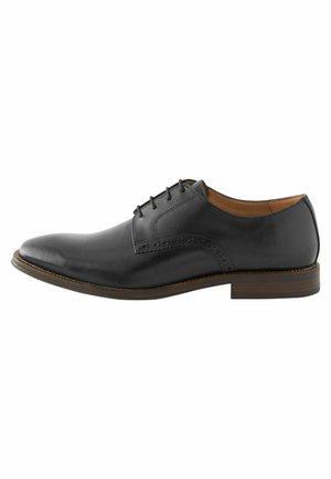 Zapato de vestir de cuero negro con punta redonda, cierre de cordones y detalles de diseño perforados. Presenta un tacón apilado marrón y una textura suave.
