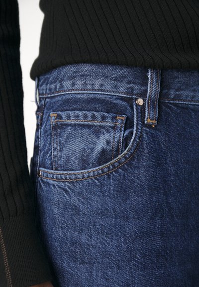 Un jeans en denim bleu présente une coupe classique, avec une poche arrière visible et des détails de couture. Associé à un pull noir à côtes.