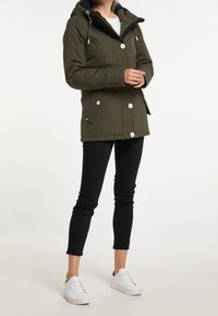 Veste imperméable vert olive avec capuche, équipée de boutons blancs, deux poches avant et des accents noirs. Portée avec un pantalon noir ajusté et des chaussures blanches.