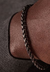 Metaal kettingarmband met in elkaar grijpende schakels, met een donkere afwerking, gedragen op de huid met een textuur, tegen een grijze achtergrond.