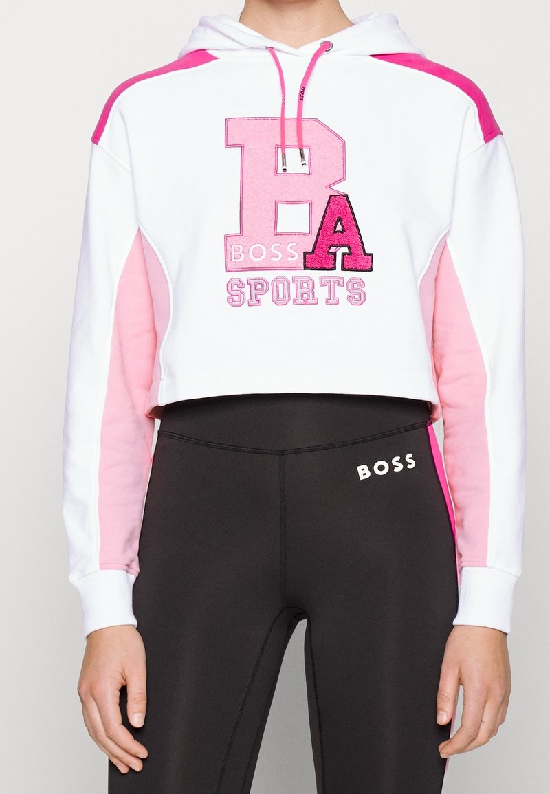 Sweat à capuche court blanc avec des blocs de couleur rose, grand lettrage "B A BOSS SPORTS" en rose et noir, porté avec des leggings noirs affichant le logo "BOSS".