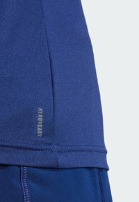 Haut de sport bleu en tissu extensible, avec un design de coutures plates et une étiquette "AEROREADY" sur l'ourlet pour la gestion de l'humidité.