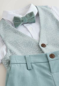 Chemise blanche avec gilet à motif floral vert clair, nœud papillon vert clair assorti et pantalon vert clair avec des boutons marron.