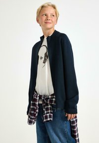 Marinblå stickad zip-up-jacka över en vit t-shirt med tryck, kombinerad med löst sittande blå jeans och en röd- och rutig skjorta knuten i midjan.