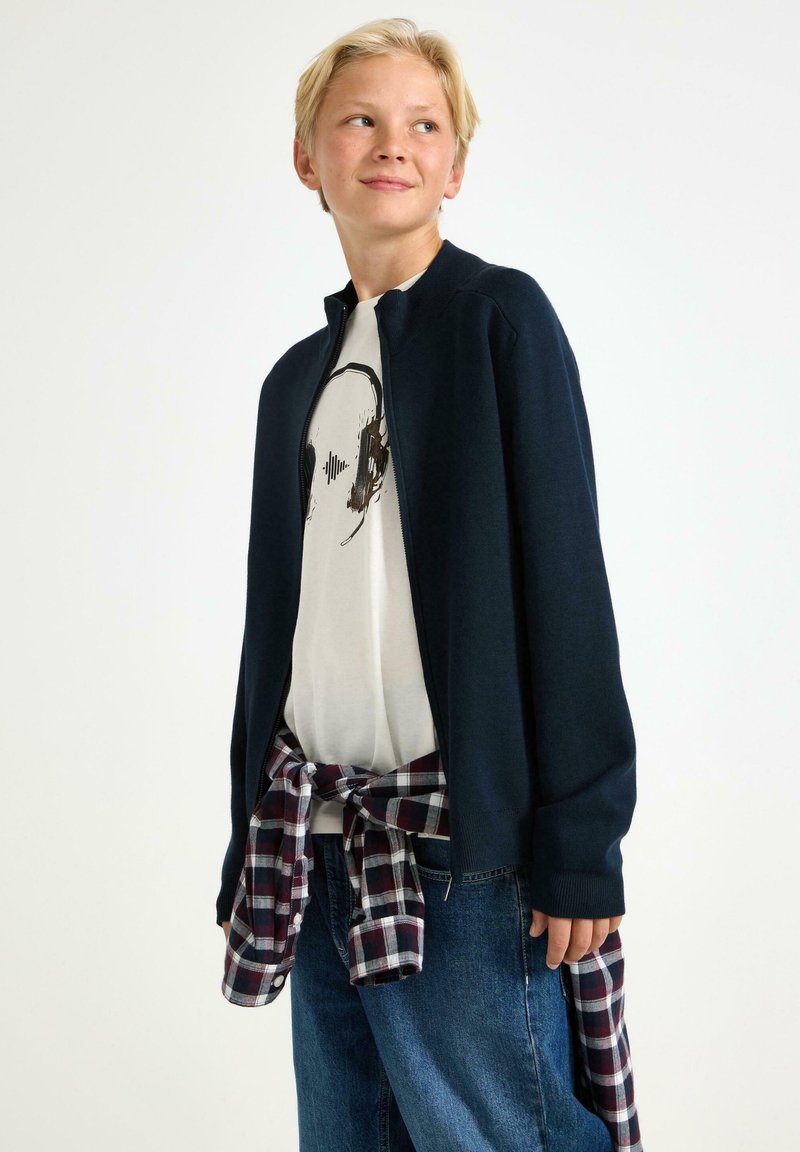 Marinblå stickad zip-up-jacka över en vit t-shirt med tryck, kombinerad med löst sittande blå jeans och en röd- och rutig skjorta knuten i midjan.