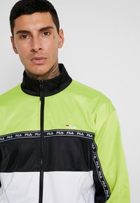 Chaqueta FILA en verde, negro y blanco con cuello alto, cierre de cremallera y diseño de bloque de color horizontal con acentos de marca.
