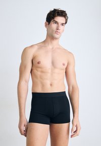 Schwarze Boxershorts mit einem Bund. Weicher Stoff, eng anliegendes Design und glatte Textur. Das Modell zeigt sie im Stehen.