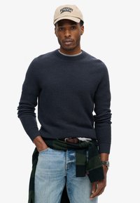 Dunkelblauer strukturierten Pullover mit Rundhalsausschnitt, kombiniert mit hellblauen Jeans. Ein grün kariertes Hemd ist um die Taille gebunden, und eine beigen Mütze sitzt auf dem Kopf.