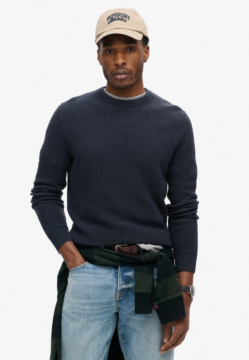 Dunkelblauer strukturierten Pullover mit Rundhalsausschnitt, kombiniert mit hellblauen Jeans. Ein grün kariertes Hemd ist um die Taille gebunden, und eine beigen Mütze sitzt auf dem Kopf.