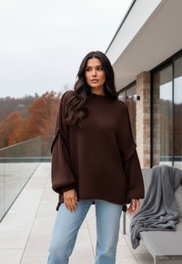 Brązowy sweter z dzianiny o wysokim kołnierzu i oversize'owych rękawach, w połączeniu z jasnoniebieskimi dżinsami. Tekstura materiału jest gruba i przytulna.
