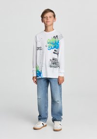 Biała koszulka z długim rękawem z kolorowymi wzorami graffiti, w połączeniu z jasnoniebieskimi zniszczonymi dżinsami i białymi trampkami z brązowymi akcentami.