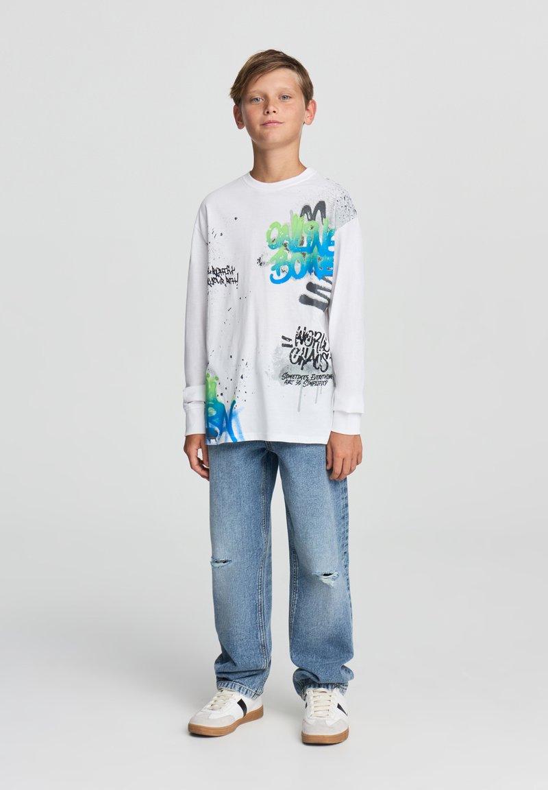 Biała koszulka z długim rękawem z kolorowymi wzorami graffiti, w połączeniu z jasnoniebieskimi zniszczonymi dżinsami i białymi trampkami z brązowymi akcentami.