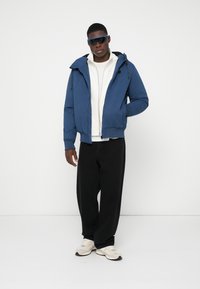 Man staat met een blauwe hooded jacket, een witte rits-sweater, zwarte ruime broek, witte sneakers en donkere zonnebril tegen een effen achtergrond.
