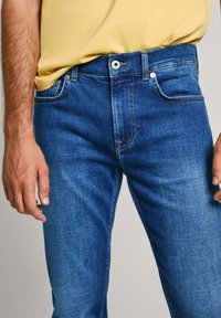 Jeans en denim bleu avec une coupe droite, léger effet délavé et détails de couture jaunes. Comprend une fermeture à bouton et zip, ainsi que des poches avant.