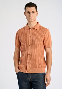 Kortärmad orange stickad piké med ett texturerat kabelmönster, knäppt framtill och klassisk krage, tillsammans med mörka jeans i denim.