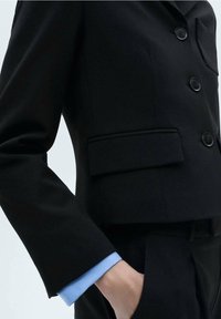 Blazer noir sur mesure avec une coupe courte, doté de boutons, d'une poche latérale et d'un détail bleu aux poignets. Tissu lisse avec une coupe structurée.