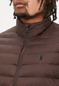 Brun puffjacka med horisontell sömnad, hög krage och svart dragkedja. Har en liten svart logotyp på bröstet. Mjuk, slät textur.