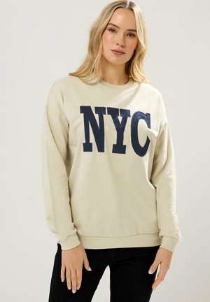 Blonde Frau trägt einen beigen Pullover mit großen marineblauen "NYC"-Buchstaben, kombiniert mit schwarzen Hosen, steht vor einem einfarbigen Hintergrund.
