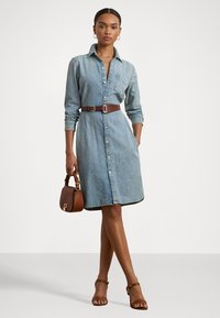 Denim shirt dress, light blue, button-front with long sleeves. Brown belt cinches waist. Brown handbag and tan heeled sandals complete the look.
