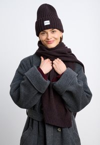 Ciemnoszary wełniany płaszcz z dużymi klapami, owijany wzorzystym bordowym szalem. Ciemnobrązowa dziergana czapka z białą metką z przodu.