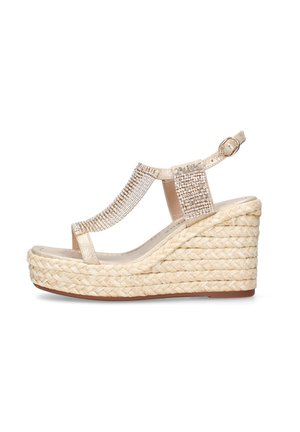 Sandalia de cuña beige con plataforma trenzada, tira en T adornada con pedrería y hebilla ajustable en el tobillo sobre fondo blanco.