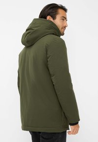 Giacca isolante verde oliva con cappuccio, caratterizzata da una superficie liscia, polsini elasticizzati e chiusura frontale con zip.